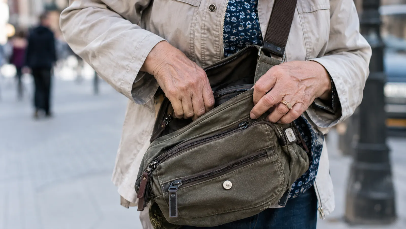 Close view of an older woman's hands checking the pockets of her crossbody messenger bag on a city street.