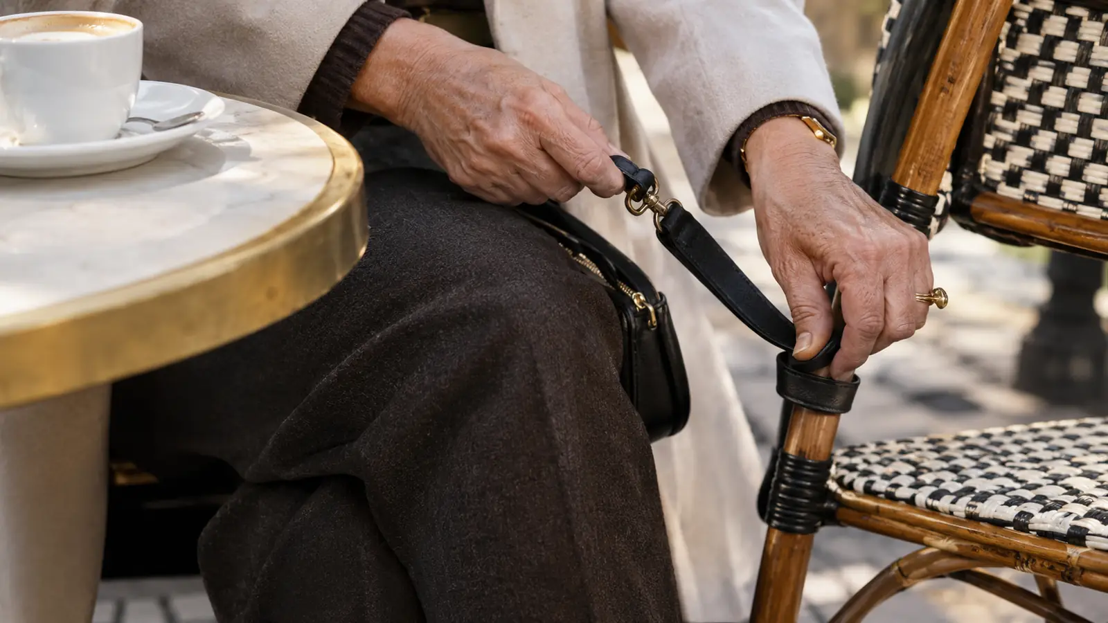 A close view of an older woman's hands unclipping the strap of a crossbody bag to secure it around a chair leg at an outdoor cafe
