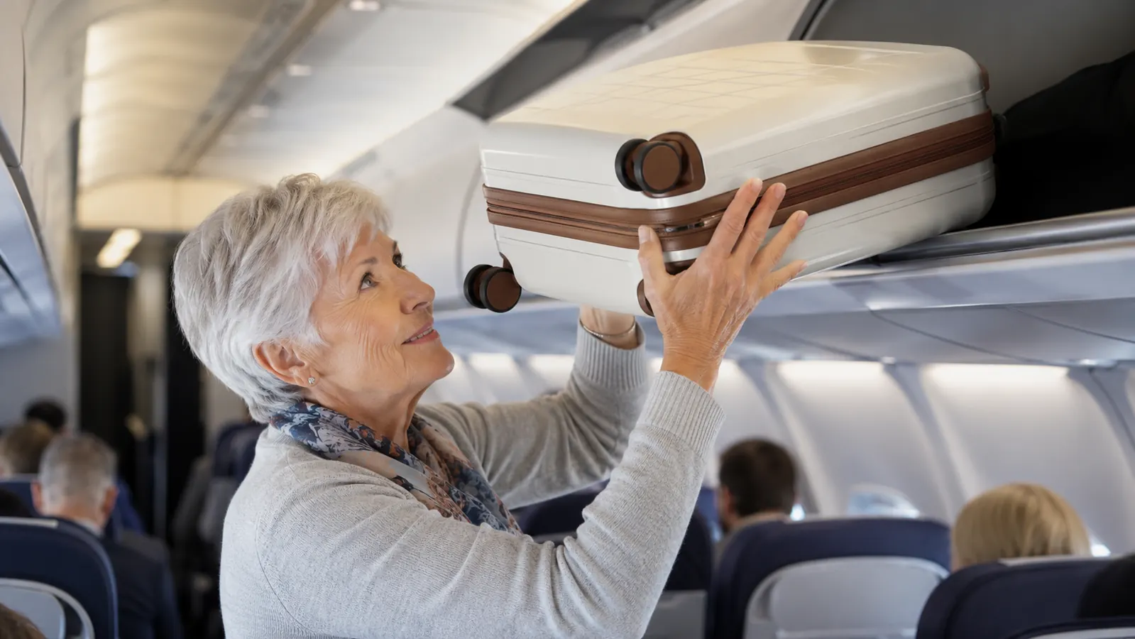 An older woman lifting a carry-on suitcase into an overhead bin on a plane